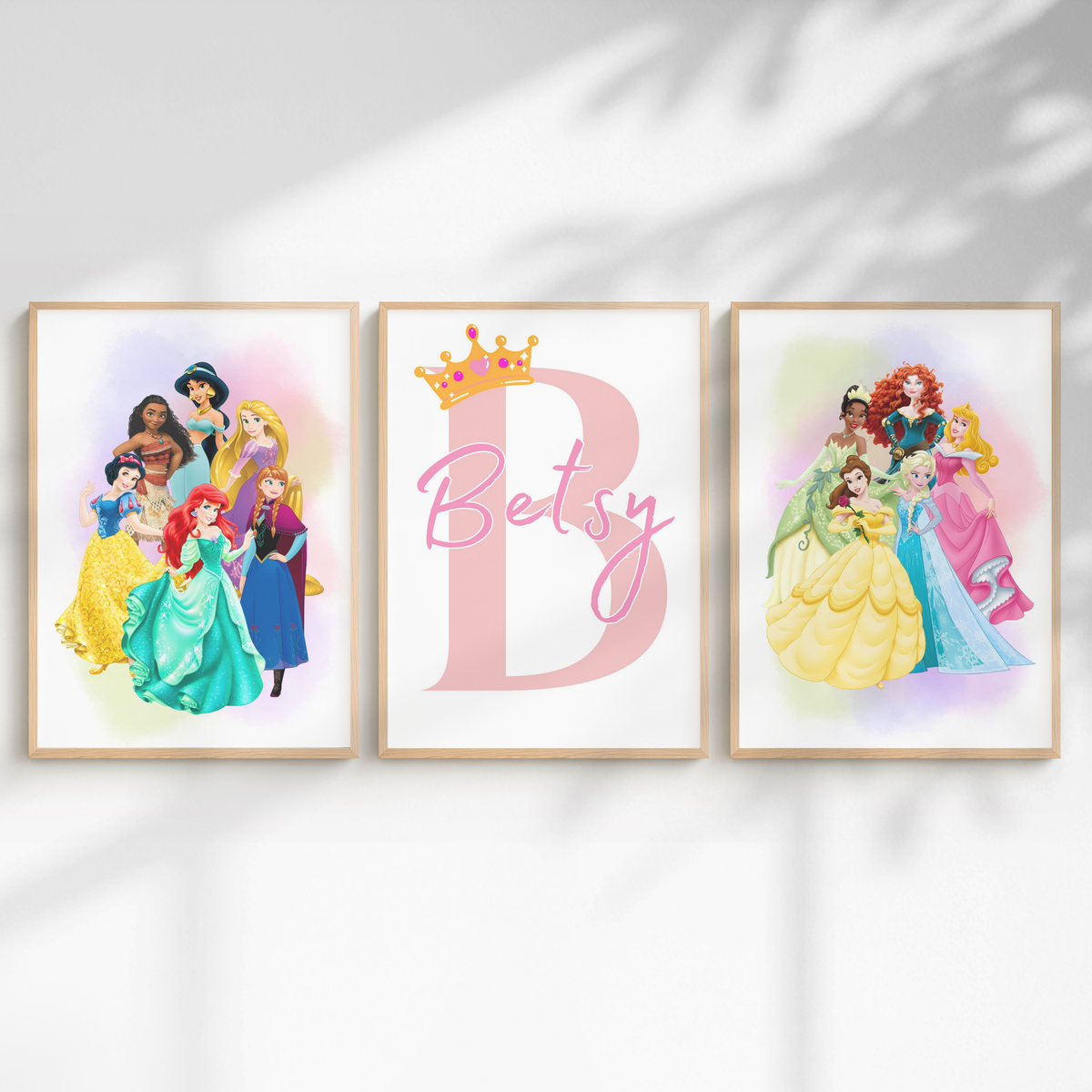 Three framed personalized wall art prints displayed side-by-side on a white wall with soft leaf shadows. The left frame shows a group of Disney princesses, the center frame features a large pink letter 'B' with a gold crown and the name 'Betsy' in script, and the right frame shows another group of Disney princesses.