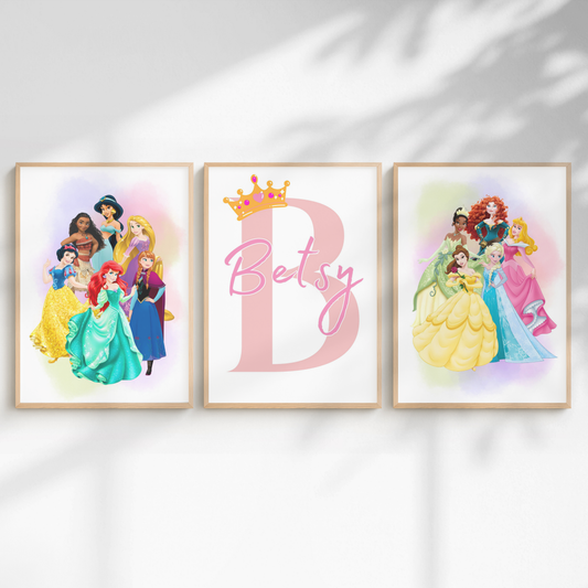 Three framed personalized wall art prints displayed side-by-side on a white wall with soft leaf shadows. The left frame shows a group of Disney princesses, the center frame features a large pink letter 'B' with a gold crown and the name 'Betsy' in script, and the right frame shows another group of Disney princesses.