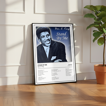 A framed 'Stand By Me' Ben E. King poster leaning against a white paneled wall on a wooden floor, with a potted plant to the right.