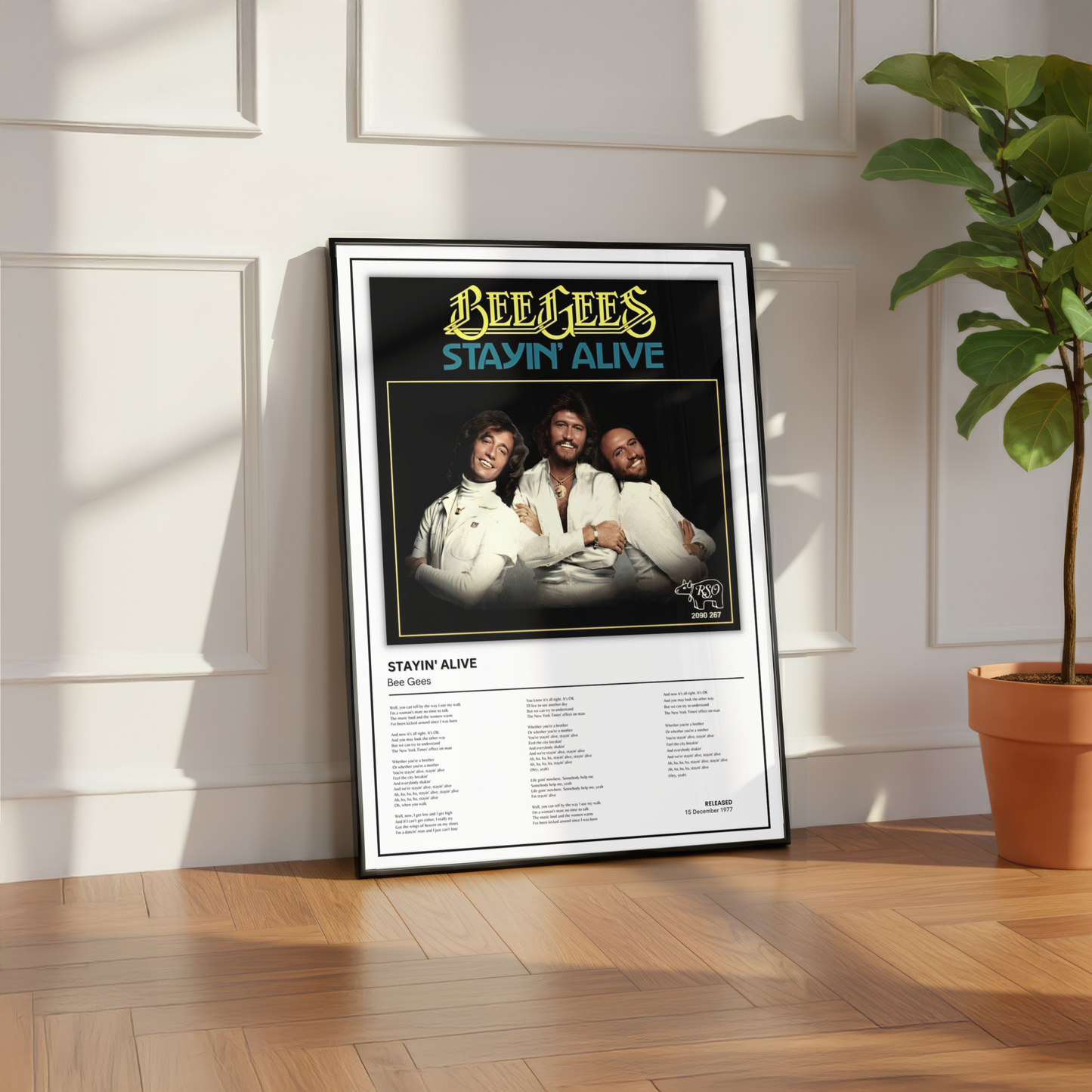 A framed Bee Gees 'Stayin Alive' music poster leaning against a white paneled wall on a wooden floor, with a potted plant to the right.