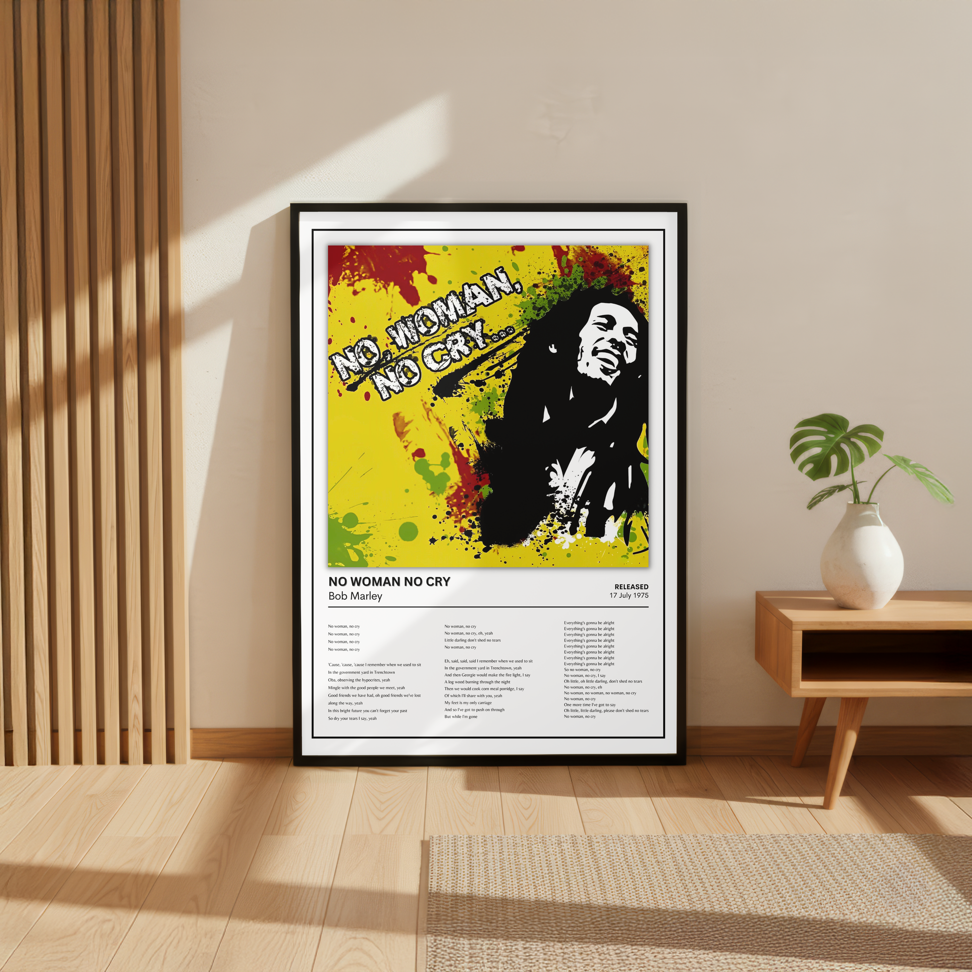 A framed Bob Marley 'No Woman, No Cry' music poster is displayed on a light beige wall in a modern room setting with a wooden slatted screen, a small wooden side table, a white vase with a green plant, and a woven rug on the floor.