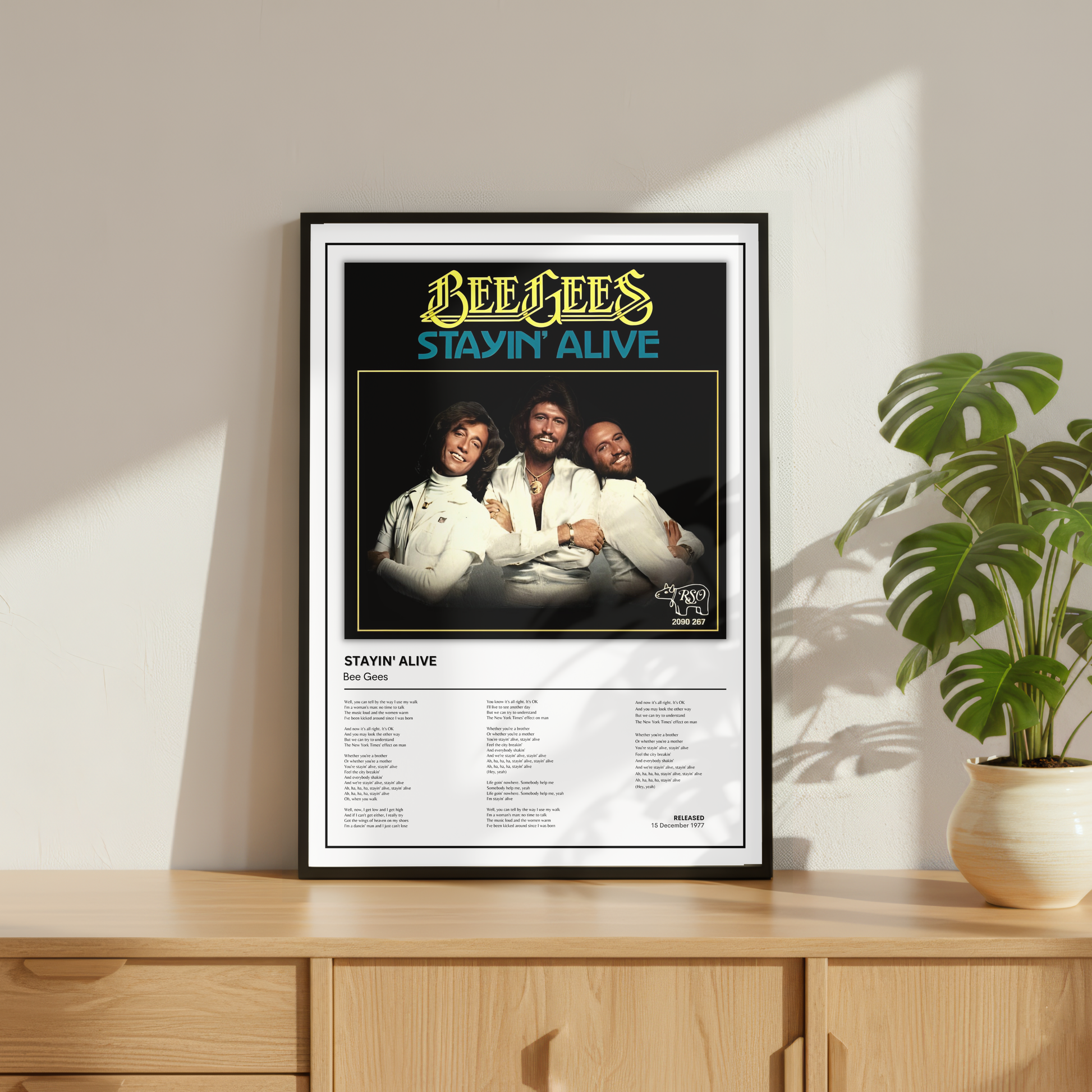 A framed Bee Gees 'Stayin' Alive' music poster displayed on a light wood console table next to a potted monstera plant, set against a warm beige wall with soft window shadows.