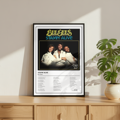 A framed Bee Gees 'Stayin' Alive' music poster displayed on a light wood console table next to a potted monstera plant, set against a warm beige wall with soft window shadows.