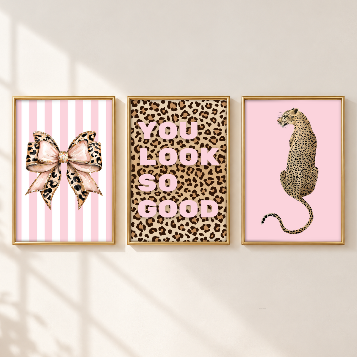 Three framed wall art prints hang side by side on a light beige wall with soft window-shadow lighting. The left print shows a pink-and-white striped background with a leopard-print bow illustration.