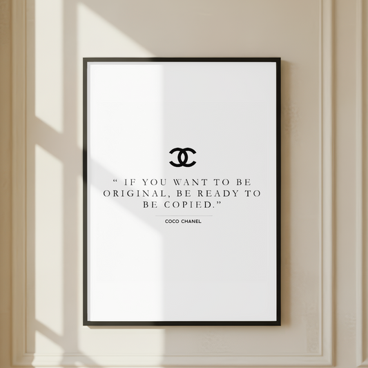 A framed wall art print with a black Chanel interlocking CC logo and the quote, 'IF YOU WANT TO BE ORIGINAL, BE READY TO BE COPIED.' by Coco Chanel, displayed on a cream-colored wall with soft window shadows.