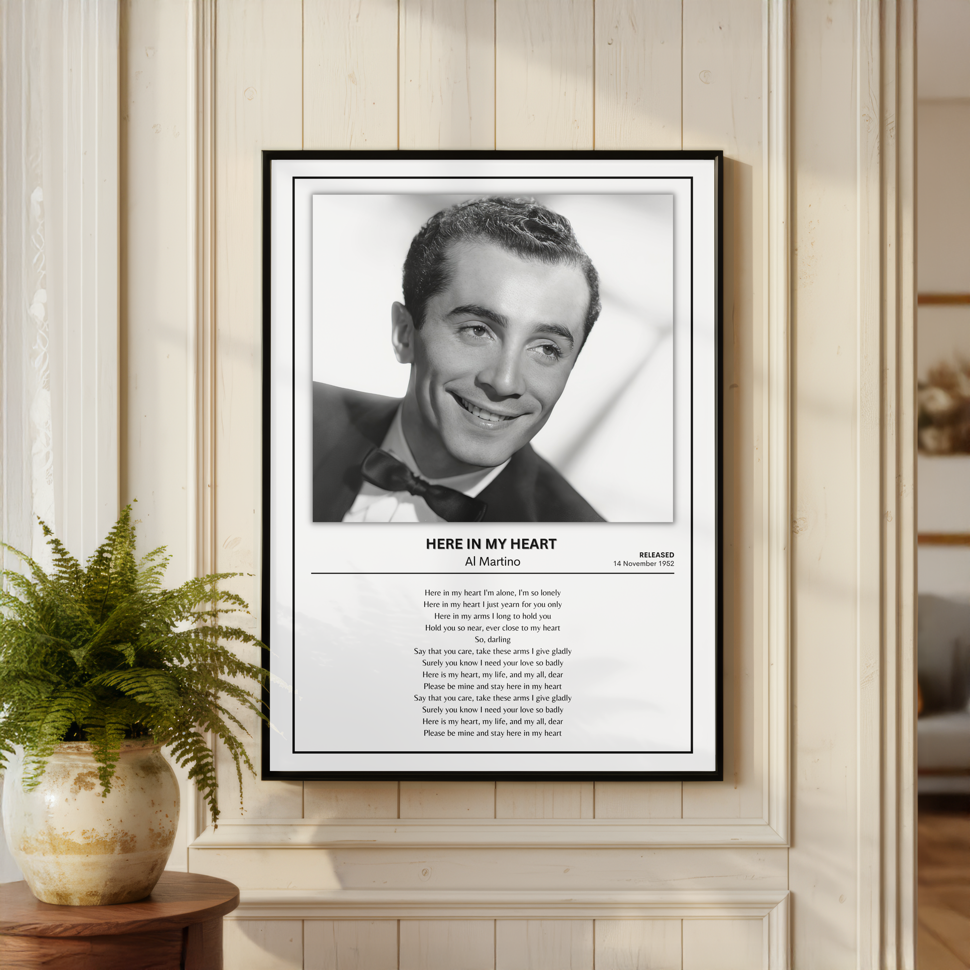 A framed black-and-white portrait print of Al Pacino with the text 'HERE IN MY HEART' and the song lyrics below, displayed on a cream paneled wall above a small table with a potted fern.