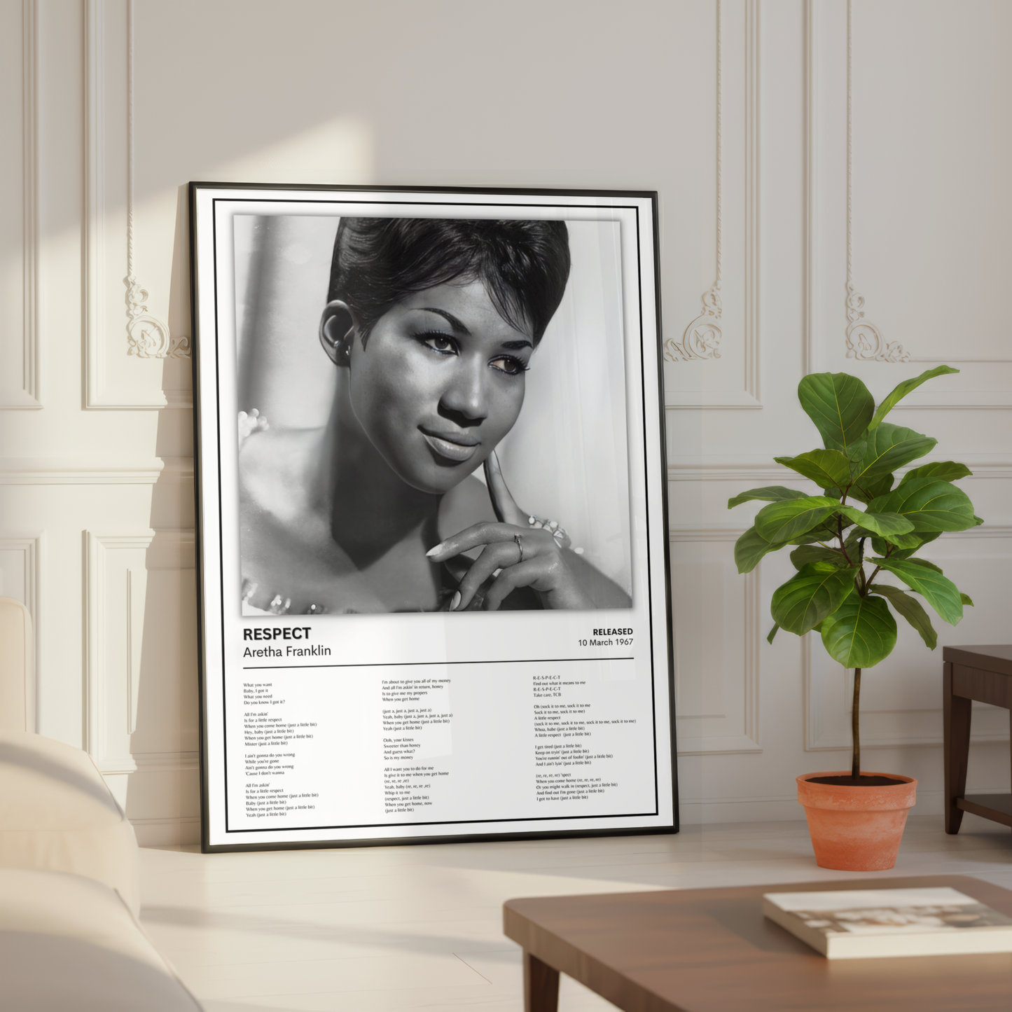 A framed black-and-white poster of Aretha Franklin leaning against a cream paneled wall in a styled room, with a potted plant and wooden table nearby.