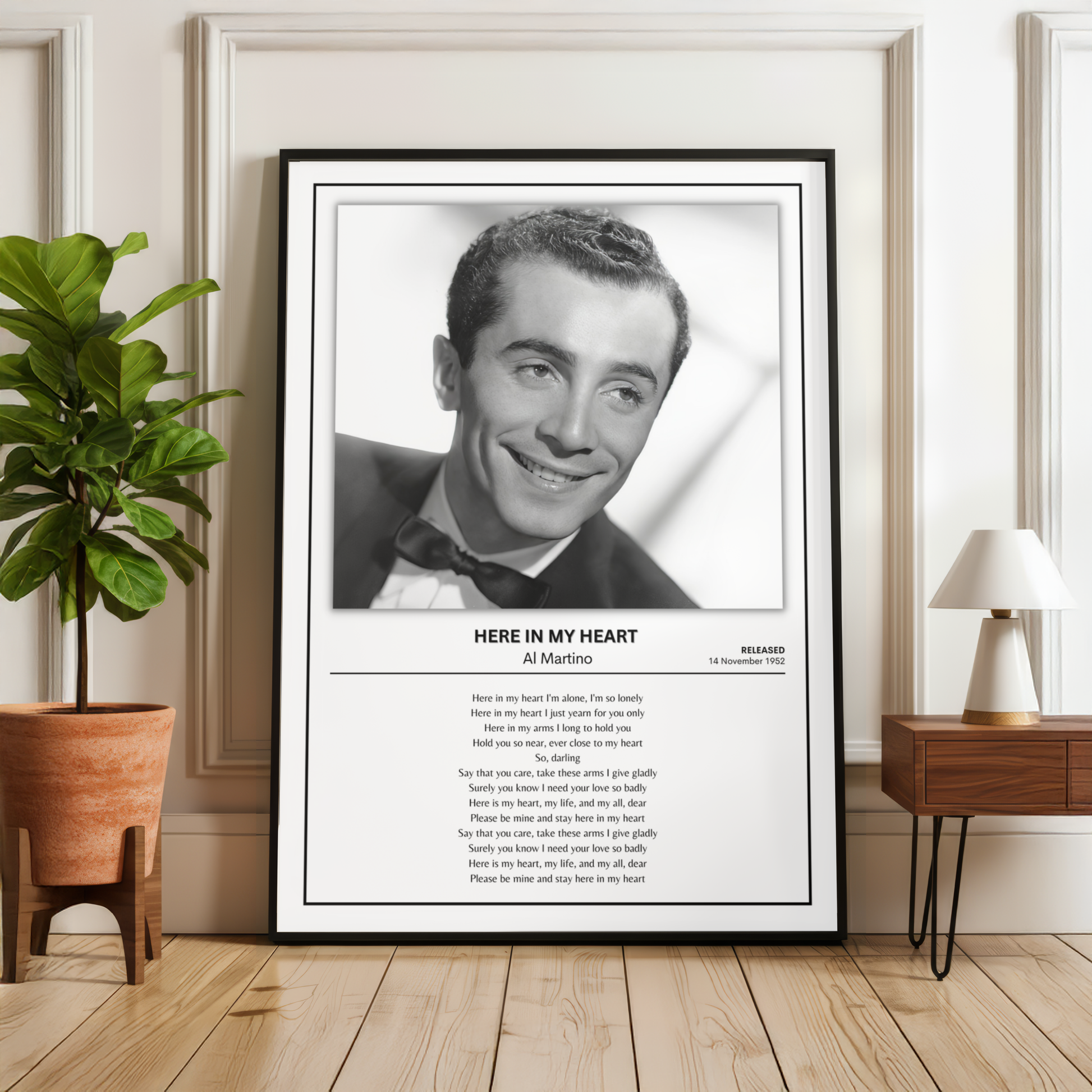 A framed black-and-white portrait print of Al Martino with the song text 'Here in My Heart' below, displayed leaning against a wall in a styled room with a plant, wood floor, and side table.