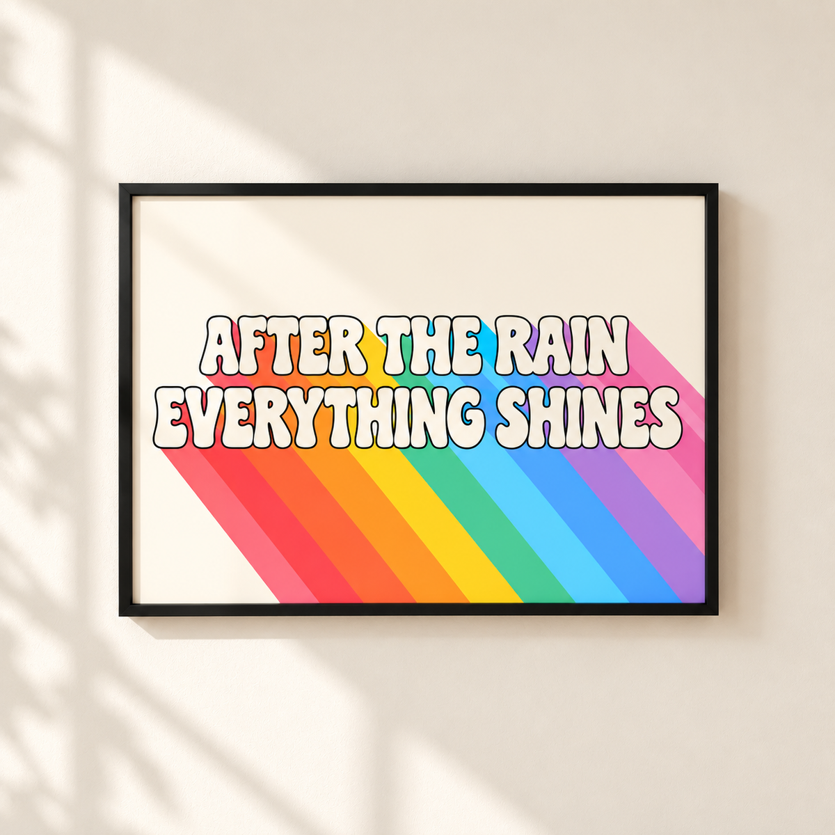 A framed art print with the text 'AFTER THE RAIN EVERYTHING SHINES' in bold white letters with a black outline, set against a retro rainbow-striped background. The print is housed in a simple black frame and mounted on a warm beige wall with soft shadow overlays.