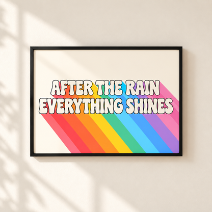 A framed art print with the text 'AFTER THE RAIN EVERYTHING SHINES' in bold white letters with a black outline, set against a retro rainbow-striped background. The print is housed in a simple black frame and mounted on a warm beige wall with soft shadow overlays.