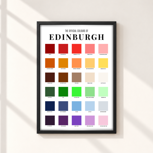 A framed Edinburgh color chart poster hanging on a light beige wall with soft diagonal shadows. The black-framed print features the text 'THE OFFICIAL COLOURS OF EDINBURGH' and a grid of colorful swatches representing the city's colors.