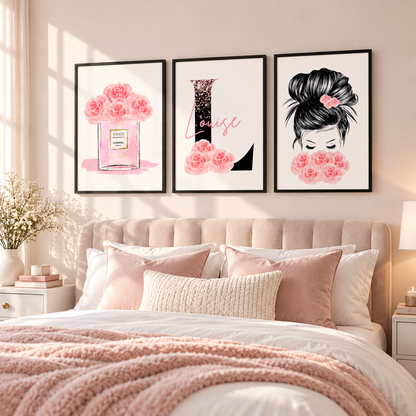 Three framed wall art prints with pink floral and elegant typography hang above a bed in a softly styled bedroom. The prints feature pink roses in a perfume bottle, the word 'Louise' in black and pink, and a black-and-white portrait with pink roses.