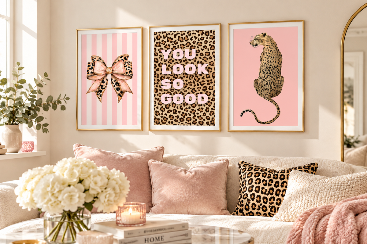 Three framed wall art prints hang above a sofa in a styled living room. The left print shows a pink bow on pink stripes, the center print says 'YOU LOOK SO GOOD' in pink text over a leopard print background, and the right print shows a leopard cat on pink.