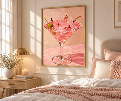 A framed art print depicting a pink martini cocktail with olives, styled on a neutral wall above a bed in a warm, modern bedroom setting.