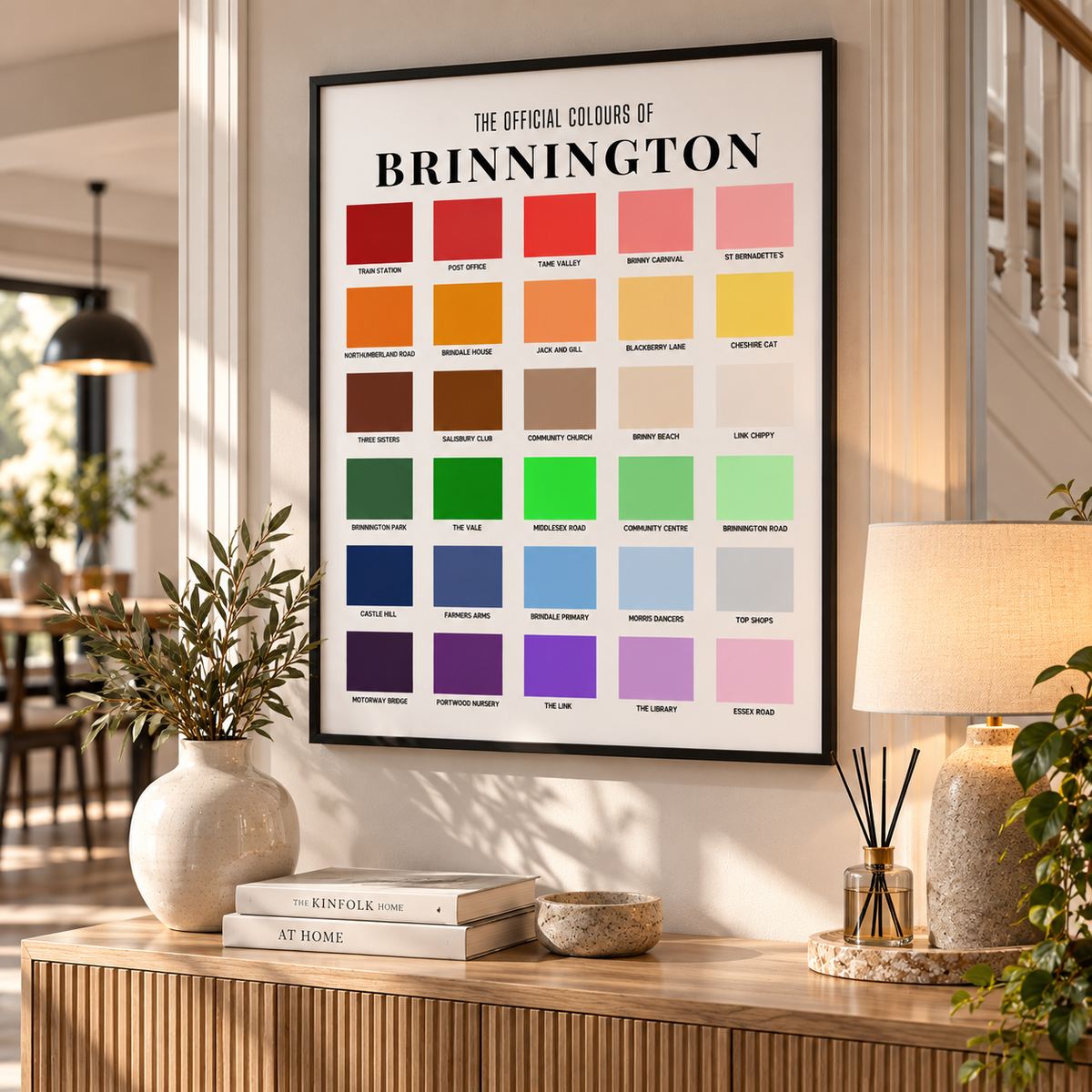 A framed 'The Official Colours of Brinnington' poster hangs on a wall in a styled home interior. The poster features a grid of color swatches with text labels, and the room includes a wooden console table, a lamp, books, and potted plants.
