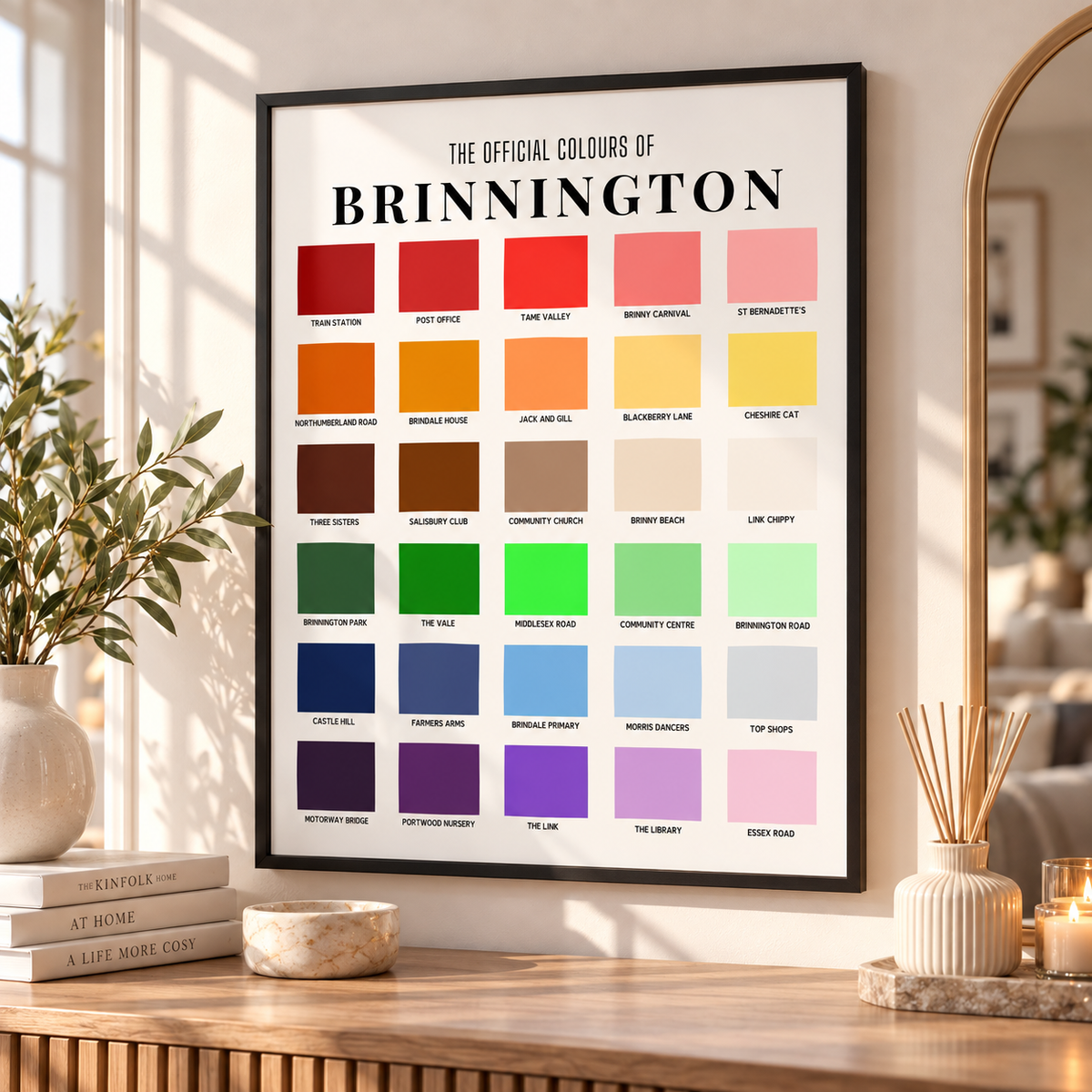 A framed wall print titled 'The Official Colours of Brinnington' hangs on a wall above a wooden table. The print shows a grid of color swatches with labels, styled in a bright, modern room with books, a vase, candles, and a mirror.