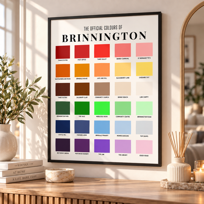 A framed wall print titled 'The Official Colours of Brinnington' hangs on a wall above a wooden table. The print shows a grid of color swatches with labels, styled in a bright, modern room with books, a vase, candles, and a mirror.