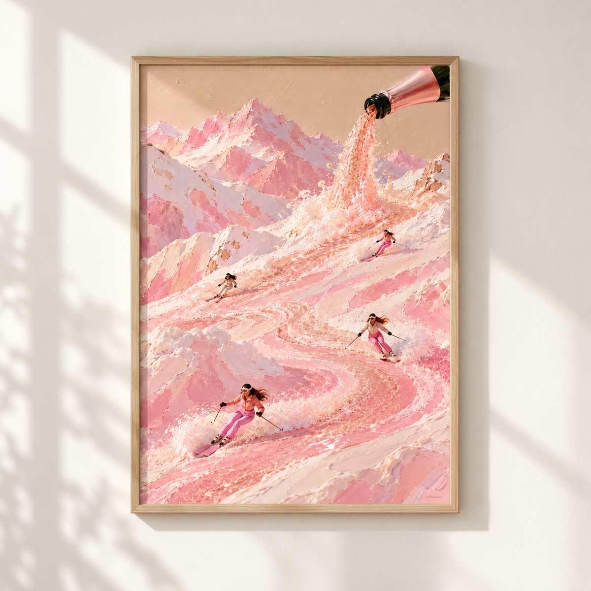 A framed art print featuring a whimsical pink mountain landscape with skiers and a champagne bottle pouring liquid, displayed on a white wall with soft window shadows.