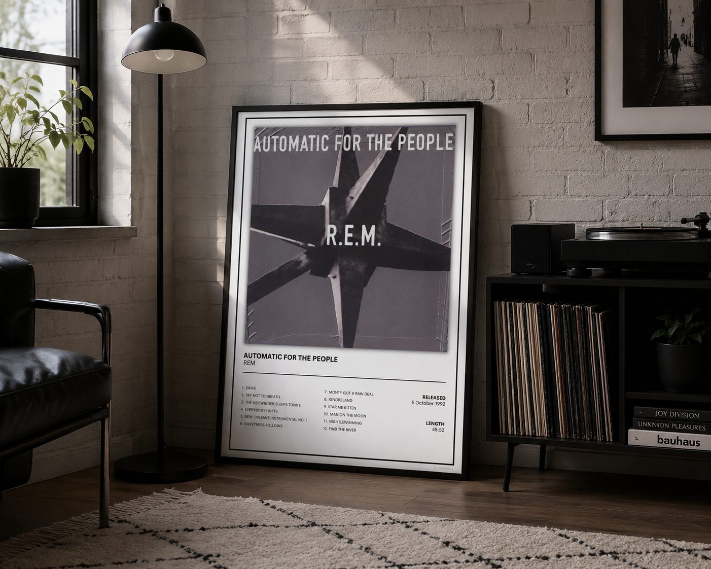 A framed black-and-white poster leaning against a white brick wall in a styled room with a floor lamp, chair, rug, and record player. The poster reads 'AUTOMATIC FOR THE PEOPLE' and features a large airplane graphic with 'A.