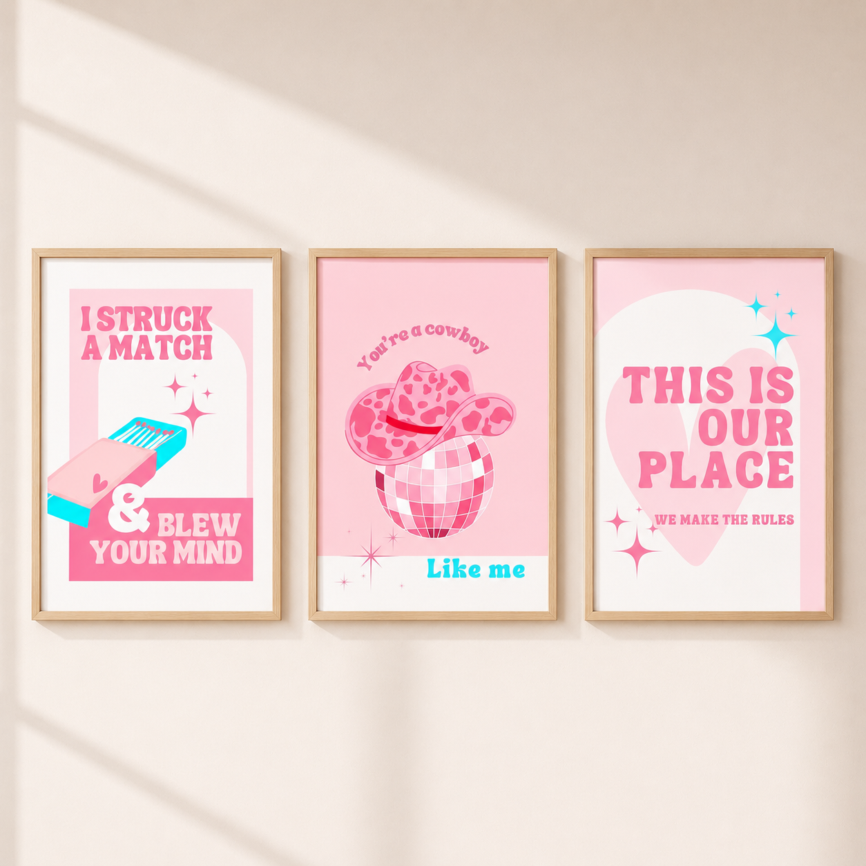 Three framed pink western-themed art prints hanging on a light beige wall. The prints feature playful text and illustrations including a match, a cowboy hat in a drink, and a heart with the phrase 'THIS IS OUR PLACE'.