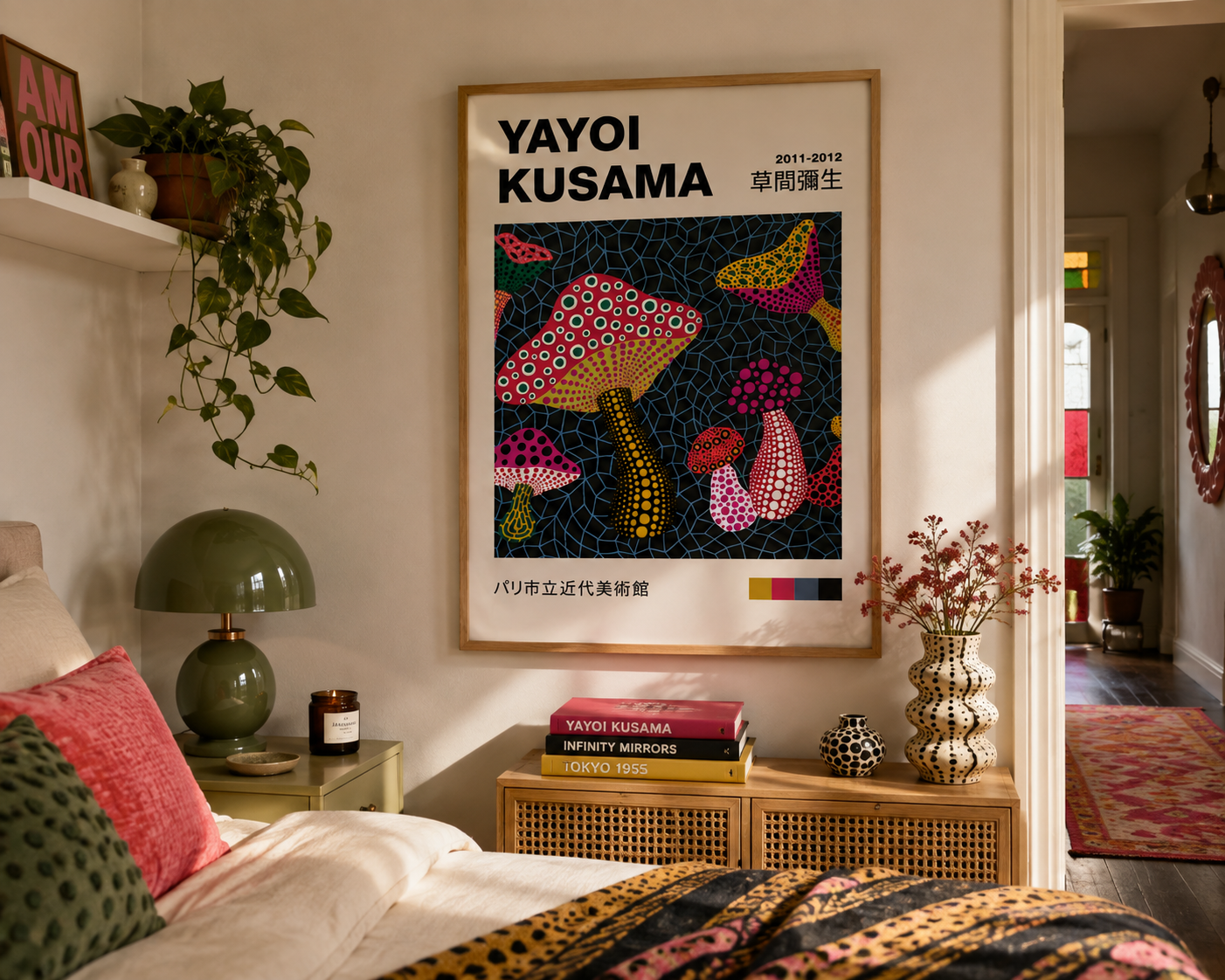 A framed Yayoi Kusama poster featuring colorful mushroom illustrations hangs on a bedroom wall above a bed. The room includes a green lamp, shelf with plants, books, and a vase, with warm natural light coming from an adjacent room.