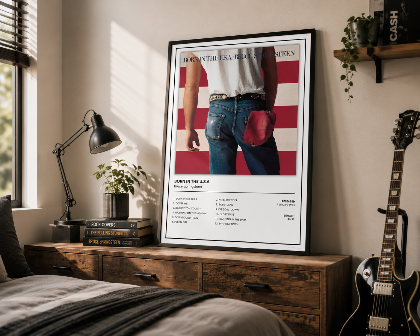 A framed poster leaning against a wall on a wooden nightstand in a styled bedroom. The poster features a vintage-style graphic with a person from behind wearing jeans and a white shirt, set against a red and white striped background, with text including 'BORN IN THE U.S.A