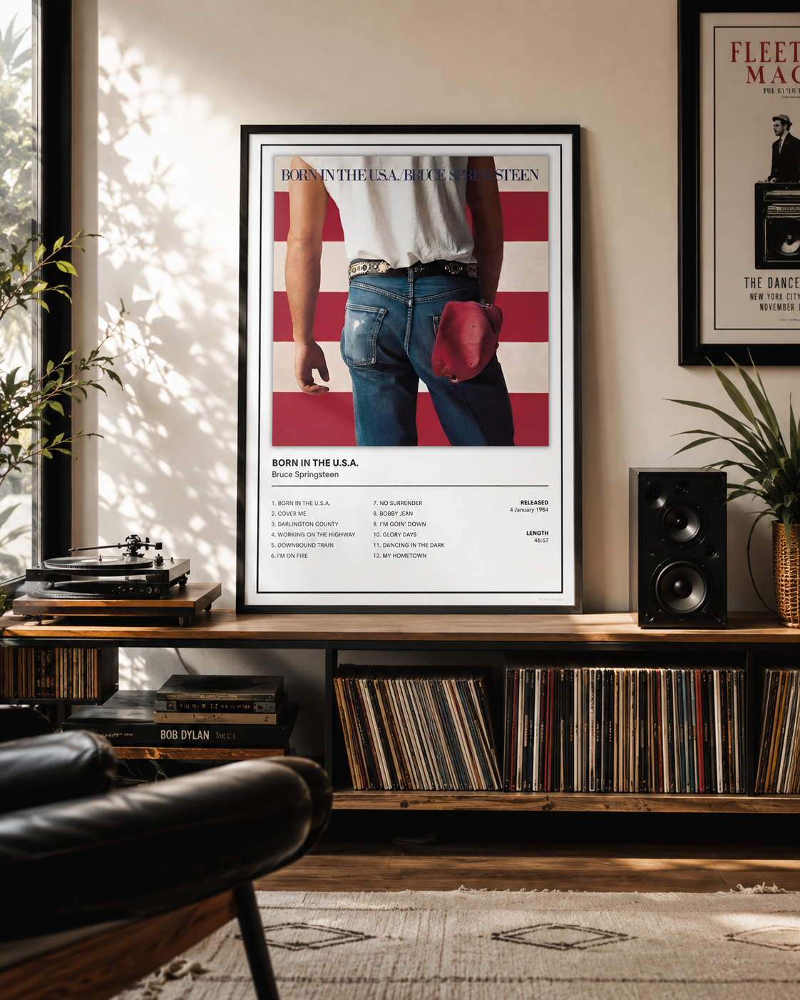 A framed music poster for Bruce Springsteen's 'Born in the USA' hangs on a wall in a styled living room with a record shelf, speakers, and plants.