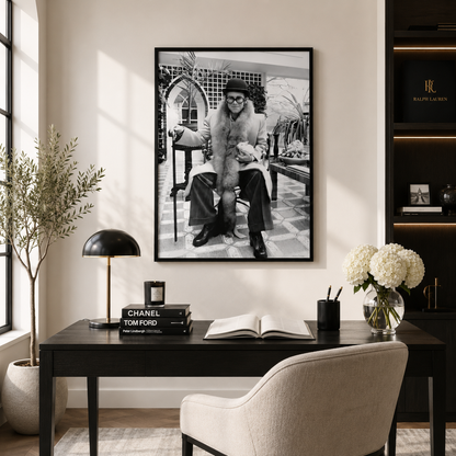 A large black and white framed art print of a vintage fashion portrait hangs on a beige wall above a dark wood desk with a chair, lamp, and decorative items.