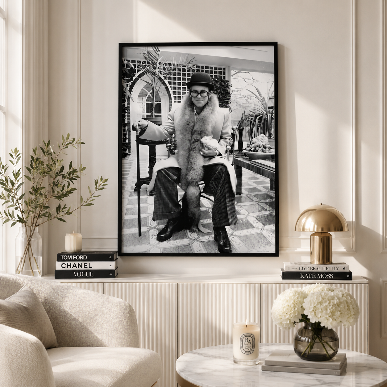 A large black and white framed art print depicting Elton John in a fur coat, styled in a modern, elegant living room setting with a beige armchair, a marble coffee table, and decorative books.
