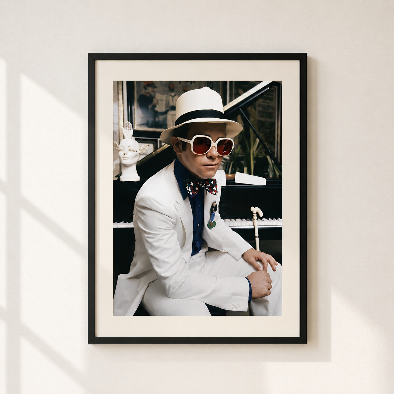 A framed art print featuring Elton John in a white suit, white fedora, white sunglasses, and a patterned bow tie, seated in front of a piano. The print is framed in black with a white mat, hanging on a light beige wall with soft window shadows.