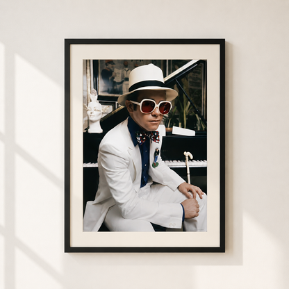 A framed art print featuring Elton John in a white suit, white fedora, white sunglasses, and a patterned bow tie, seated in front of a piano. The print is framed in black with a white mat, hanging on a light beige wall with soft window shadows.