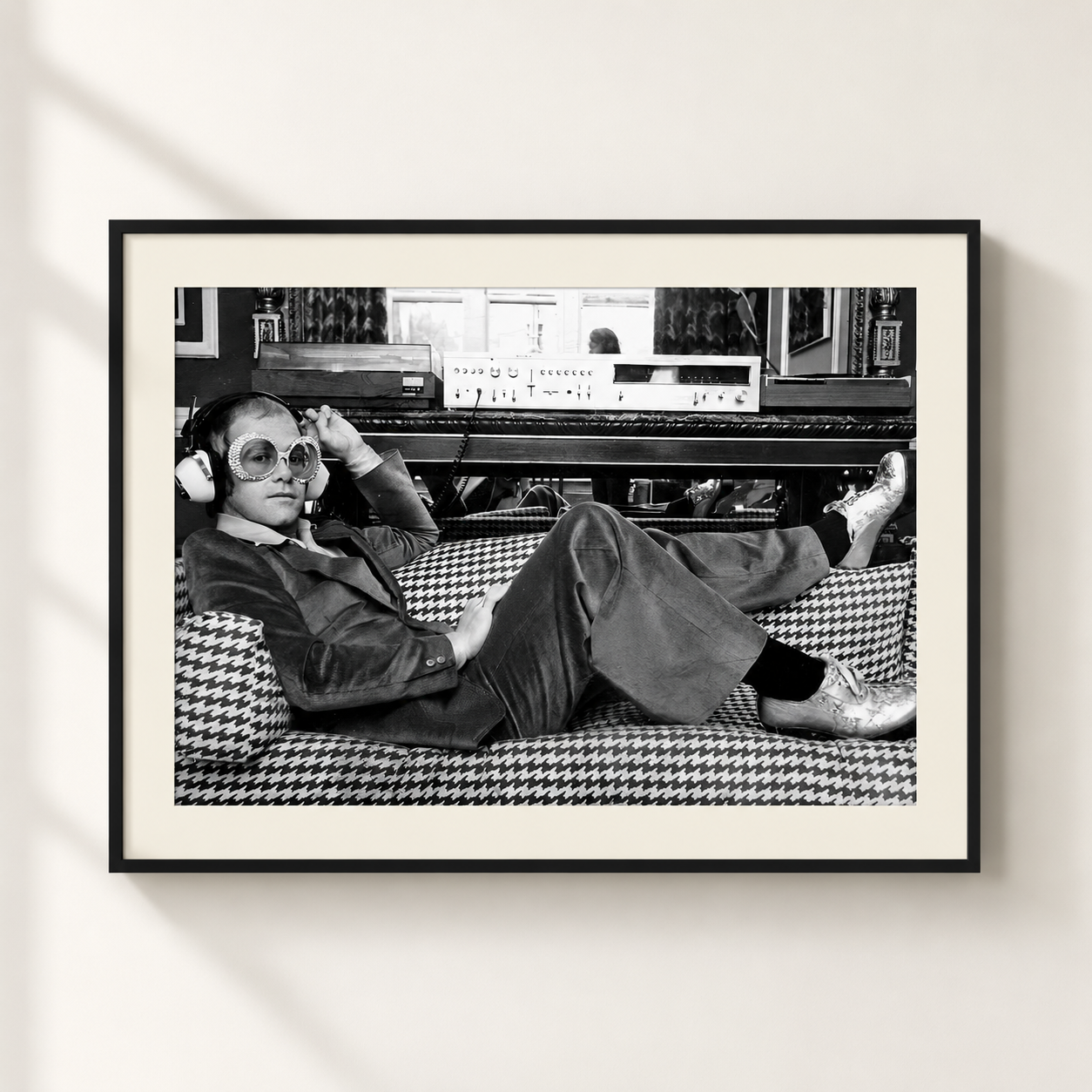 A framed black and white art print featuring a Elton John in a houndstooth-patterned suit lounging on a couch in front of audio equipment, displayed on a light beige wall.