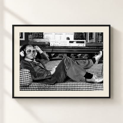 A framed black and white art print featuring a Elton John in a houndstooth-patterned suit lounging on a couch in front of audio equipment, displayed on a light beige wall.