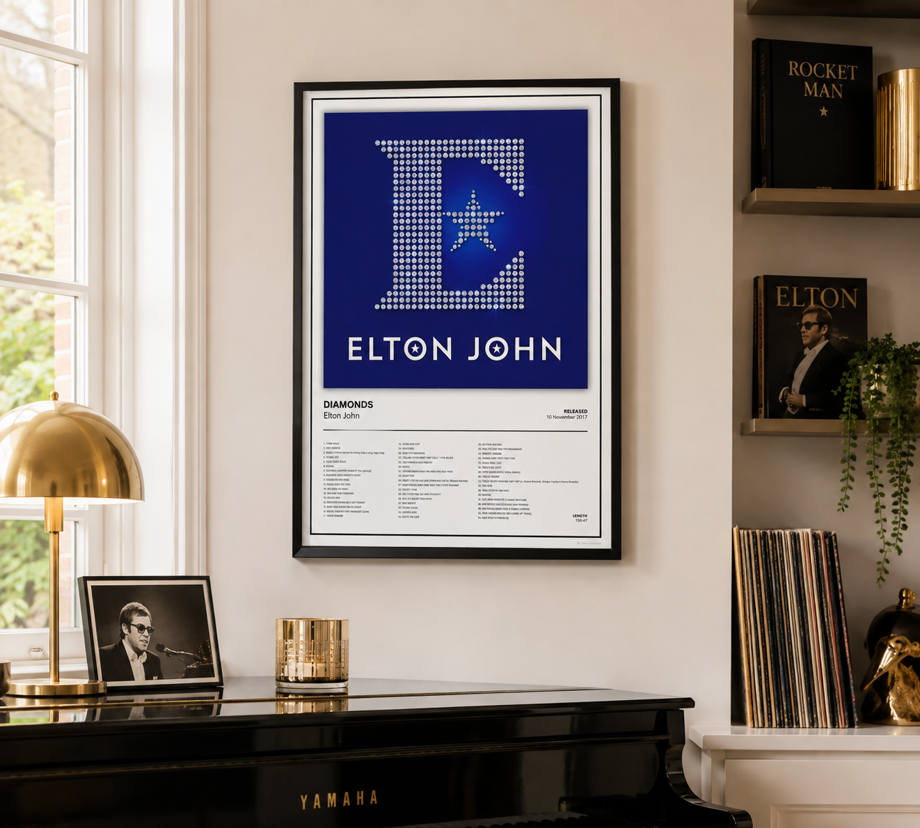 A framed Elton John 'Diamonds' music poster hangs on a cream-colored wall in a styled room setting above a black piano, with a brass lamp, records, and shelves in the background.