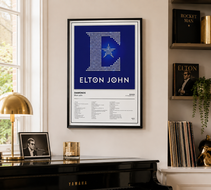 A framed Elton John 'Diamonds' music poster hangs on a cream-colored wall in a styled room setting above a black piano, with a brass lamp, records, and shelves in the background.