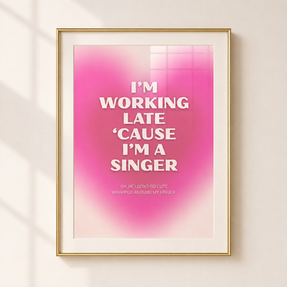 A framed typographic wall art print with a pink gradient background and white text reading, 'I'M WORKING LATE 'CAUSE I'M A SINGER,' shown hanging on a light beige wall with soft window shadows.