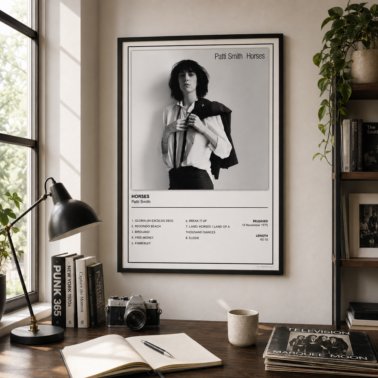 A framed black-and-white music poster of Patsy Smith Horses hangs on a beige wall in a styled room with a desk, lamp, books, camera, mug, and shelving.