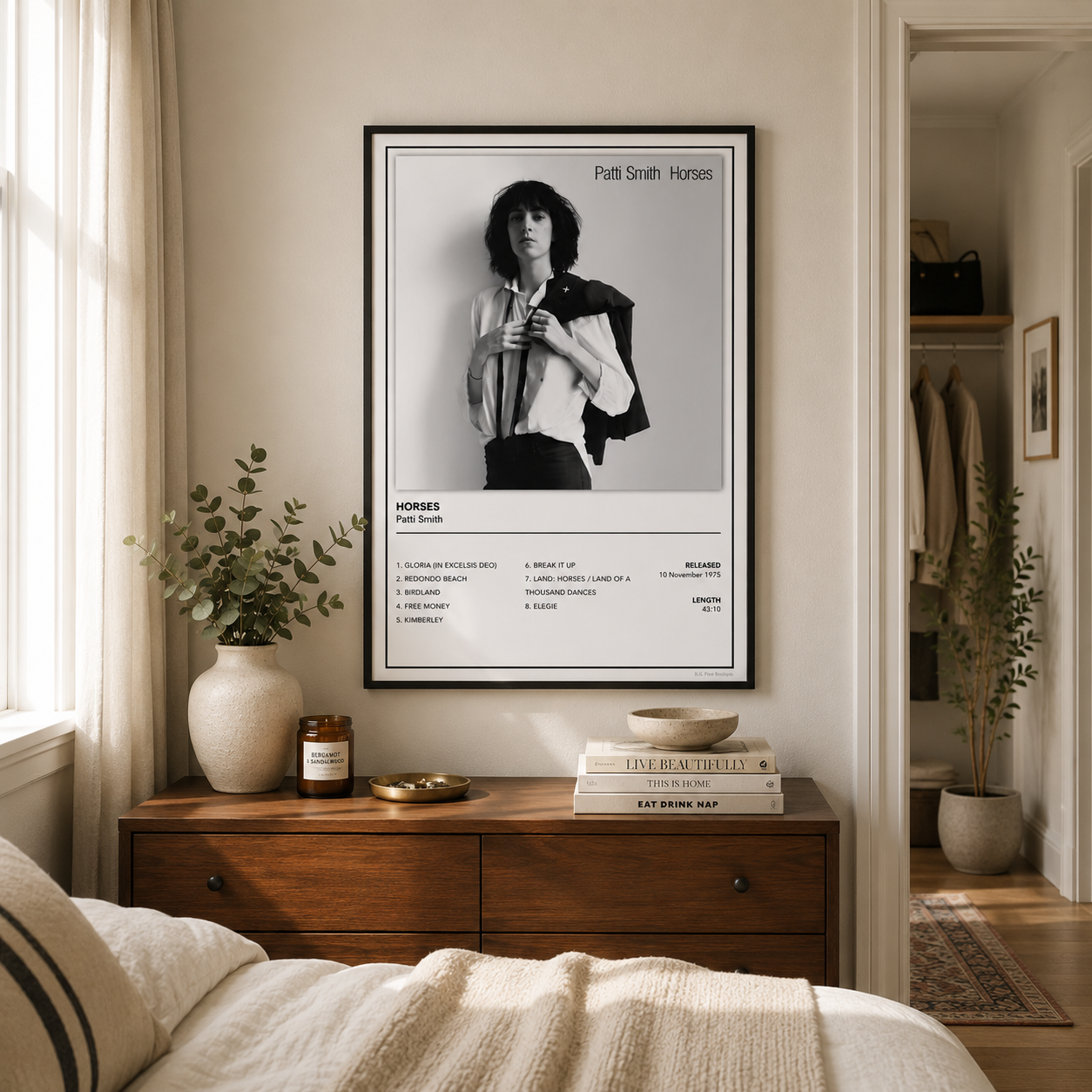 A framed black-and-white poster of Pattie Smith Horses hanging on a beige wall above a wooden dresser in a styled bedroom. The poster features a black-and-white portrait of a person with a jacket and text at the bottom.