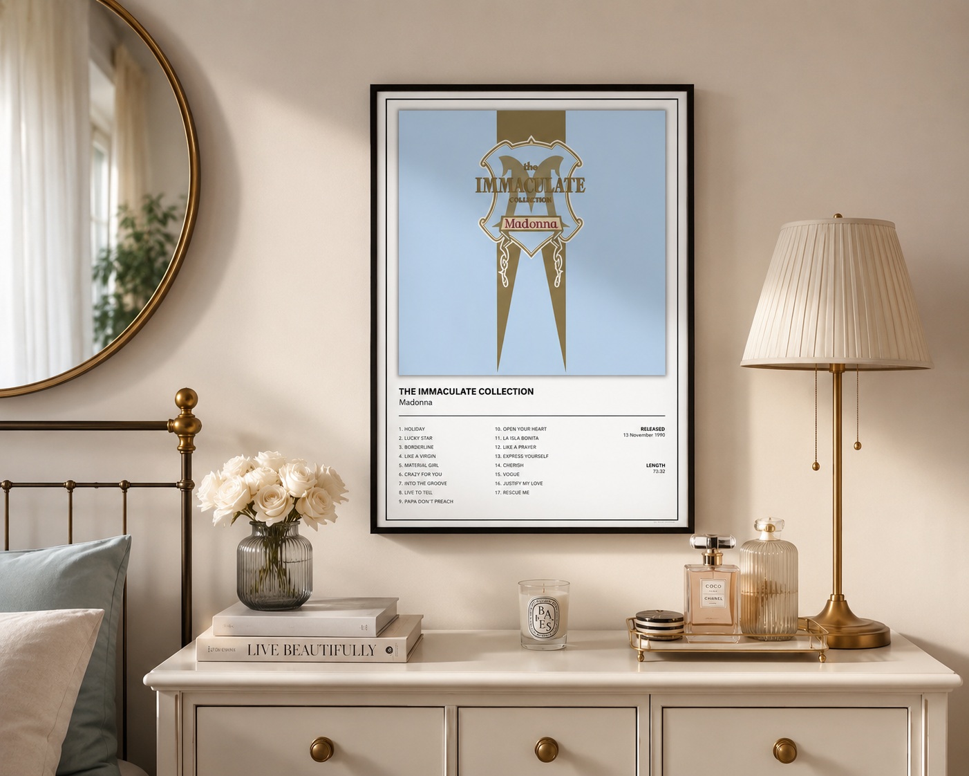 A framed wall art print featuring a stylized album-cover design with the text 'Madonna The Immaculate Collection' and track listing text below, displayed in a styled bedroom setting above a dresser with a lamp, flowers, and books.