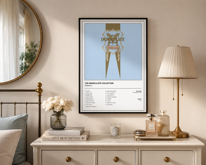 A framed wall art print featuring a stylized album-cover design with the text 'Madonna The Immaculate Collection' and track listing text below, displayed in a styled bedroom setting above a dresser with a lamp, flowers, and books.
