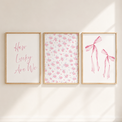 Three framed pink-themed nursery art prints hanging on a cream wall above a light wood herringbone floor. The left print has pink script text reading 'How Lucky Are We', the center has a delicate pink floral pattern, and the right has two pink ribbon illustrations.