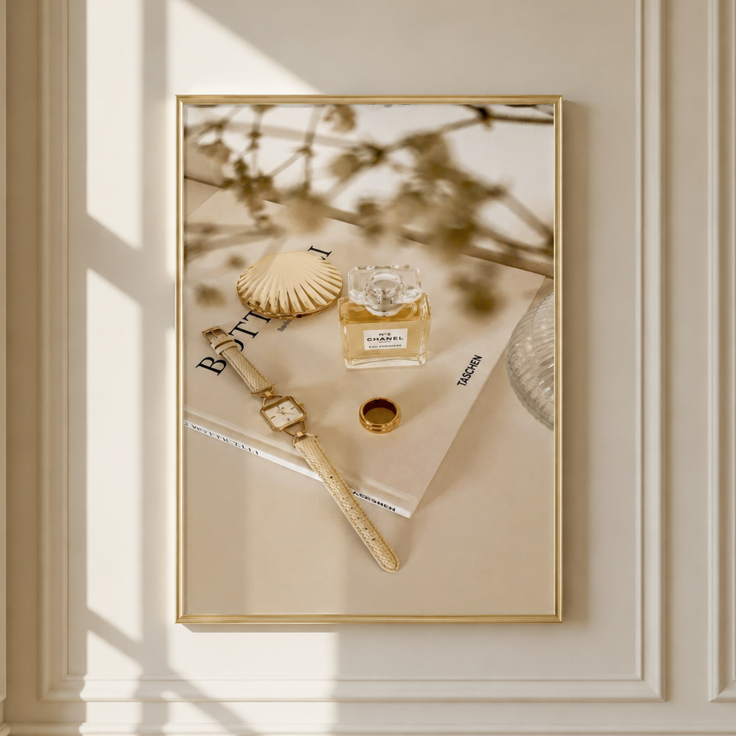 A framed art print with a gold border hangs on a cream-colored paneled wall. The print features a styled flat-lay scene with a gold watch, a seashell, a perfume bottle, and a magazine under warm, sunlit window shadows.