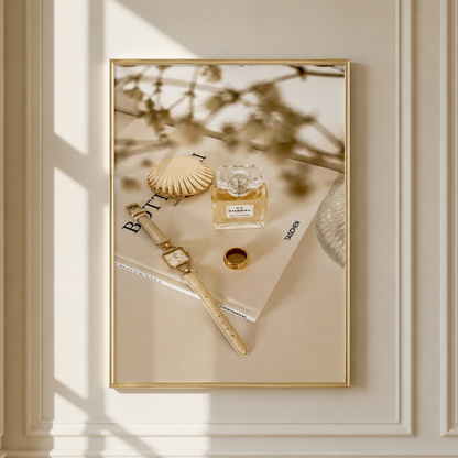 A framed art print with a gold border hangs on a cream-colored paneled wall. The print features a styled flat-lay scene with a gold watch, a seashell, a perfume bottle, and a magazine under warm, sunlit window shadows.