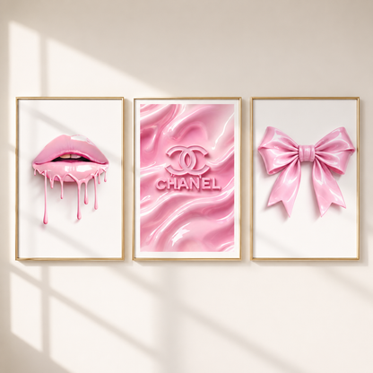 Three framed pink art prints hanging on a wall: dripping pink lips, a pink Chanel logo on a glossy pink background, and a pink satin bow. The frames are gold and the wall is light beige with soft window-shadow lighting.