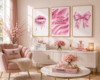 A styled pink interior scene with three framed wall art prints above a white console: dripping pink lips, a pink Chanel logo on satin, and a pink bow. A blush pink armchair with a Chanel pillow, flowers, perfume bottles, candles, and a round coffee table are arranged in the room.