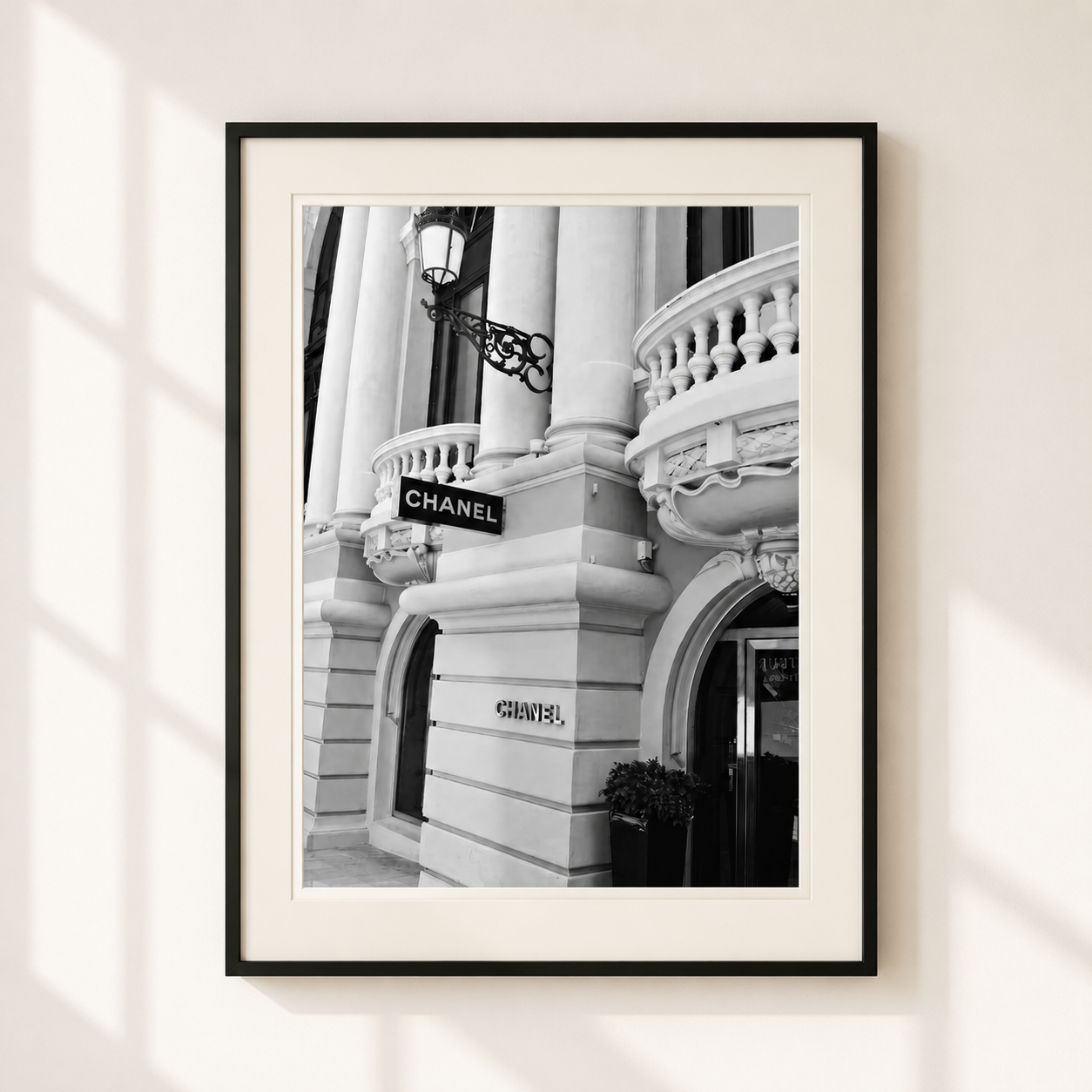 A framed black and white photograph of a classic building facade with visible CHANEL signage, displayed on a light beige wall with soft window shadow overlays.