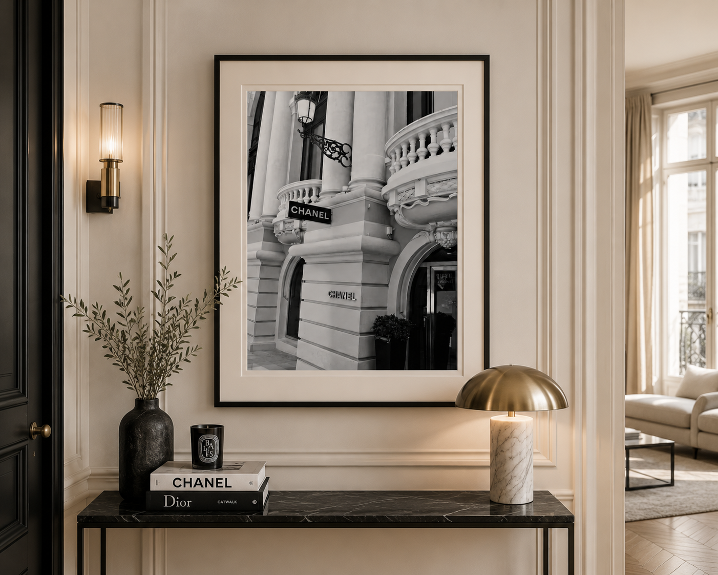 A framed black and white architectural art print featuring a building facade with a Chanel sign, displayed on a cream-colored wall above a console table decorated with a vase, books, and a lamp.