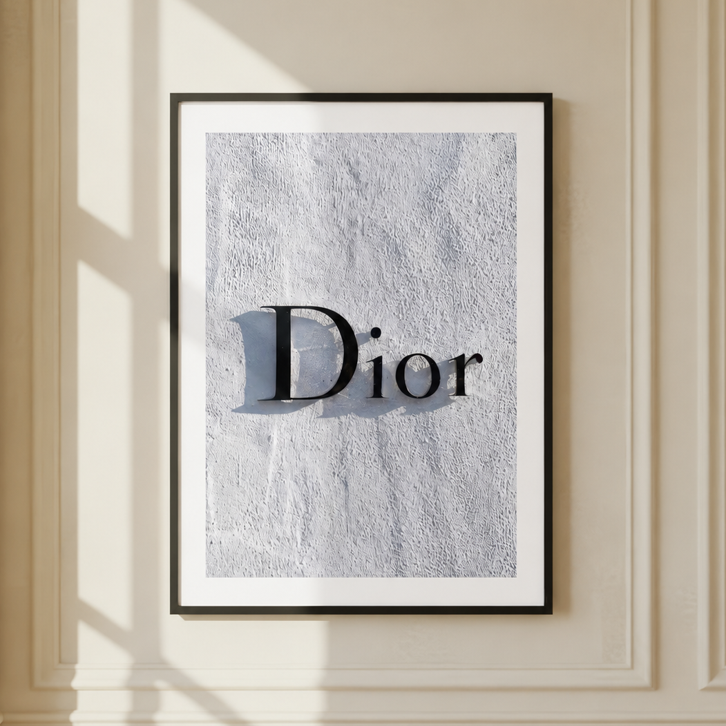 A framed art print featuring the word 'Dior' in black serif lettering over a light gray, textured background, mounted on a cream-colored wall with decorative molding and soft window shadows.