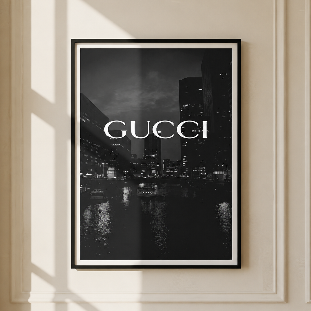 A framed black and white Gucci poster featuring a city skyline at night with the brand name in white text, displayed on a cream paneled wall with soft window shadows.