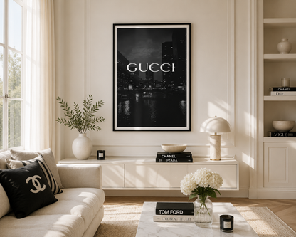 A framed black and white Gucci cityscape poster hangs on a cream wall in a styled living room with a sofa, console table, lamp, and decorative books.